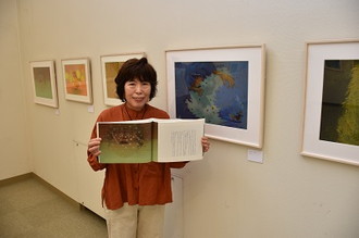 写真：作品展の図録を手に、自身の作品の前に立つ中山尚子さん