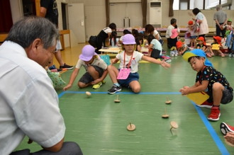 写真：床にしゃがみ込み真剣にこまを回す子どもたちと、嬉しそうに見守るお年寄り