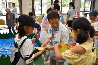 写真：首にかけたスタンプカードを手に、お年寄りに遊びの場所を聞く二人の女の子